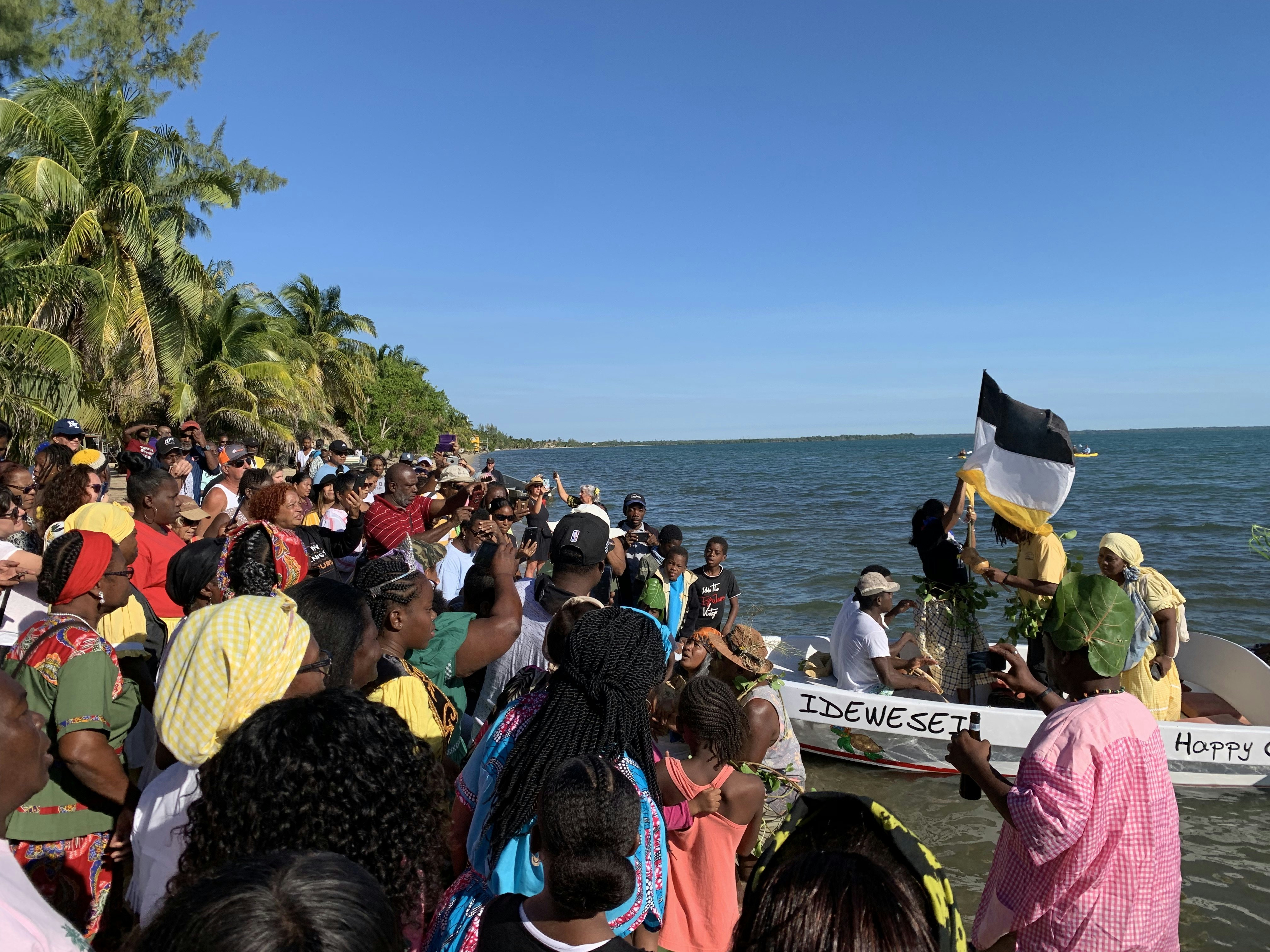 A large group of people stand on the shore watching a pair of boats filled with Garifuna actors re-enact Garifuna Settle Day.